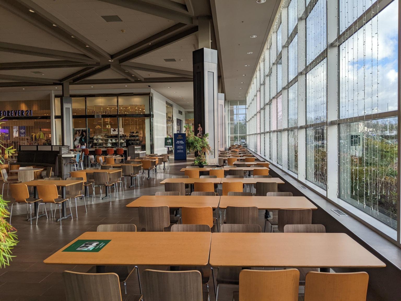 Food court inside Bellis Fair mall with rows of tables and chairs beside a wall of tall picture windows